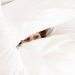 A person lying in bed mostly hidden under white blankets, with only their eyes visible, peeking out from the covers, symbolizing revenge bedtime and ADHD habits.
